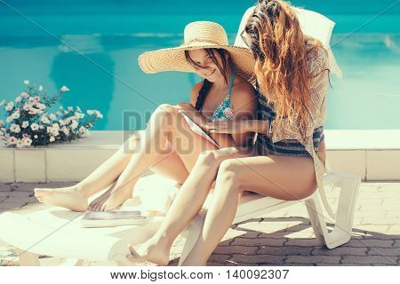 young pretty woman in glasses near small girl in straw hat reading book at swimming pool with blue water sunny summer vacation
