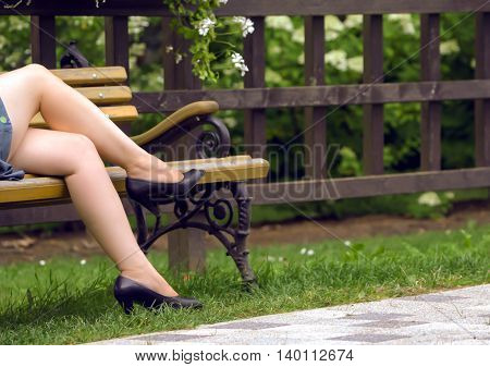 female legs in black sandals in the summer, on the bench, next to a fence with flowers, a woman short dress, legs crossed, blue dress and brown with yellow bench with armrests carved