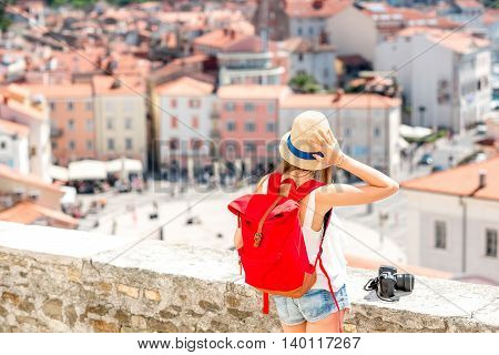 Young female traveler with hat, red backpack and photo camera enjoying the view on Piran old town. Back view with copy space. Traveling in Slovenia