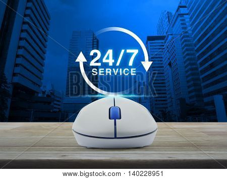 Wireless computer mouse with button 24 hours service icon on wooden table over city tower background Full time service concept