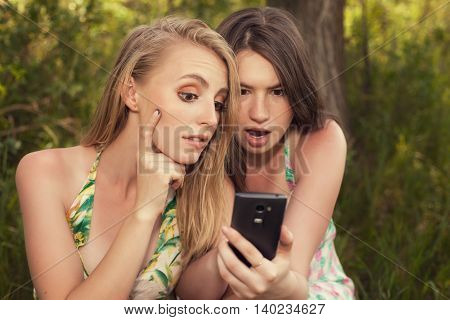 Two girls look at the phone's screen outdoors blonde and brunette is surprised