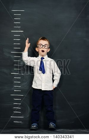 Four year old boy in white shirt and a tie standing by a stadiometer, drawn on a blackboard. Education.