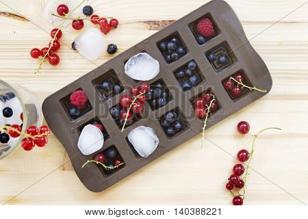 Brown ice cube tray with fresh wild berries on a wooden table: redcurrants blueberries and raspberries.