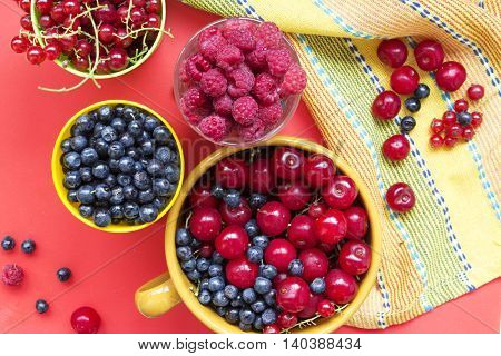 Summer fresh berries: blueberries raspberries cherries and redcurrent in bowls. Top view on a red and yellow background.