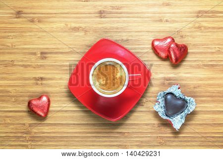 Heart shaped sweets wrapped in a bright red foil lying on a wooden texture with red cup of coffe near it. Background for romantic themes.