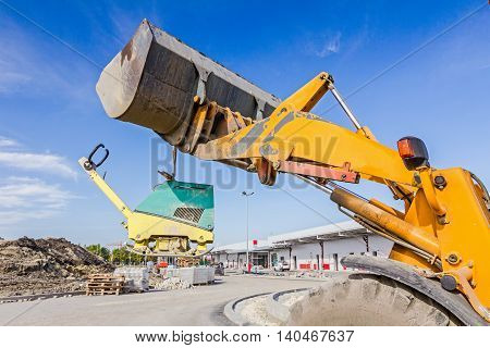 Excavator is moving over building site with raised up front bucket vibration plate compactor machine is hanging.