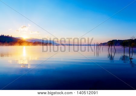 beautiful lake view in morning fog with mystic mountains and trees as leftovers of a mole in gold, purple - blue tones