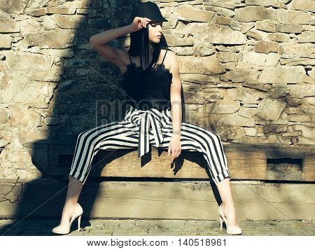 Young beautiful woman in stylish striped trausers and black hat outdoor sitting near stony wall