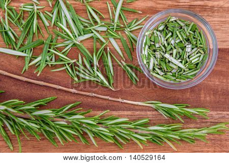 Sprigs of rosemary, and stripped and chopped rosemary (in a glass bowl) on a wood cutting board