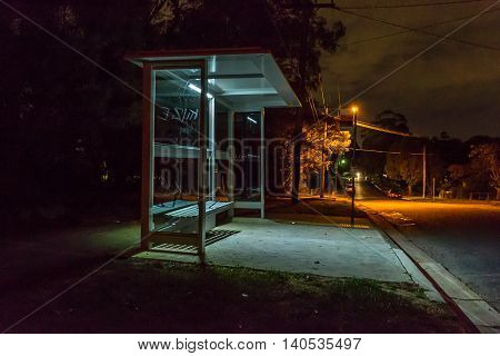 Empty bus shelter at night. Cars are parked down the road. graffiti is scratched into the glass.