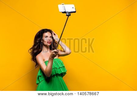 Portrait of young model in green dress and sunglasses posing while making selfie against yellow background.