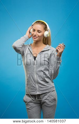 Portrait of young blonde woman in sportswear listening to music with cell phone.Studio shot. Blue background. Isolated.