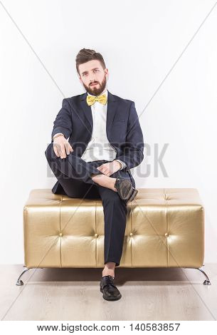 Portrait of handsome man relaxing on modern pouffe in elegant suit with bow-tie