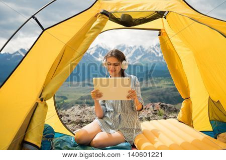 girl in the tent with headphones listening to music and looking at Tablet on mountain background