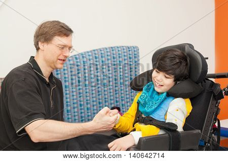 Father playing with disabled biracial son sitting in wheelchair while waiting in doctor's office laughing together.