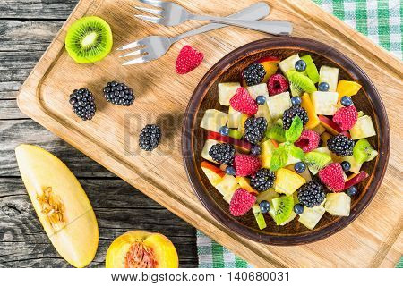 delicious fruit and berry summer salad decorated with mint leaves on clay dish on cutting board with dessert forks half of peach and slice of melon on rustic boards view from above