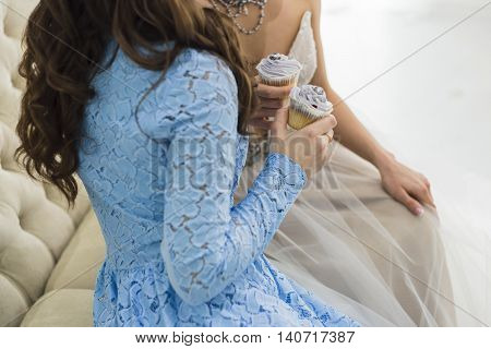 Bride in wedding dress with bridesmaid holding a beautiful blue wedding cupcakes and sitting on sofa