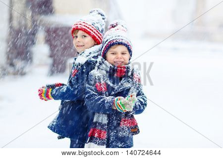Two little kid boys in colorful clothes, outdoors during snowfall. Active outoors leisure with children in winter on cold snowy days. Happy siblings having fun with snow