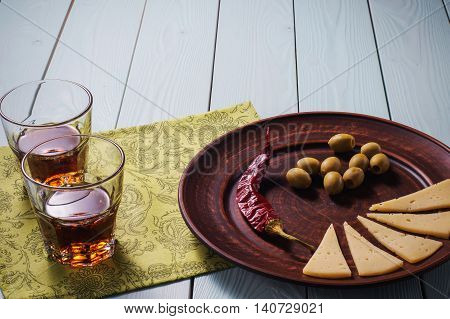 Two whiskey glasses and a plate of olives cheese and pepper on a blue wooden background.