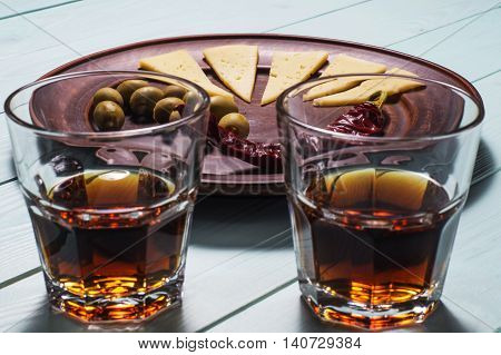 Two whiskey glasses and a plate of olives cheese and pepper on a blue wooden background.