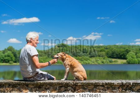 Senior man with old brown dog on wall in nature landscape with lake