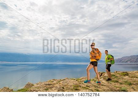 Happy couple hikers trekking in summer mountains on island seaside. Young woman and man walking with backpack on rocky mountain trail. Camping and looking at beautiful inspirational landscape view.