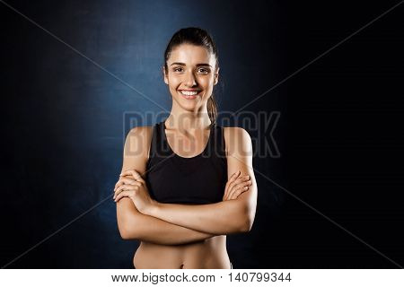 Young beautiful sportive girl posing with crossed arms over dark background. Copy space.