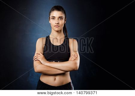Young beautiful sportive girl posing with crossed arms over dark background. Copy space.