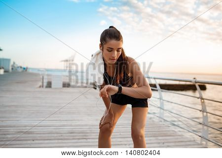 Young beautiful brunette sportive girl looking at watch at sunrise over seaside. Copy space.