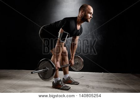 Young powerful sportsman in heaphones training with barbell over black background. Copy space.
