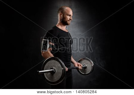 Young powerful sportsman in heaphones training with barbell over black background. Copy space.