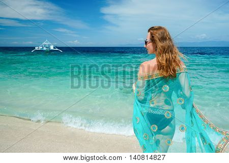 young caucasian woman with blue sari is staying on the white beach of tropical turcuoise sea at sanny day and getting plesaure under blue sky