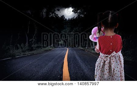 Back view of young girl is standing on a country road in a forest. Lonely child with doll sad gesture. Negative human emotion. Outdoors. The moon were NOT furnished by NASA.