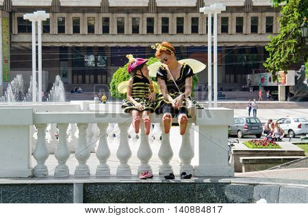 Kharkiv Ukraine - June 18 2016: Mother and daughter dreessed in costumes of bees are drying their feet after washing them in the fountain. Costumed bike ride 