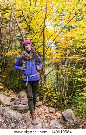 Asian woman hiking in autumn forest nature walking on trail path. Hiker girl with backpack hat and jacket on fall adventure travel outdoors relaxing enjoying good weather during hike in cold weather.