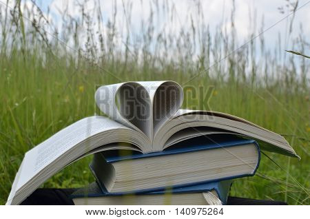 Several books on mountain meadow. The first book on the top is open and pages are forming hart figure a symbol of love.