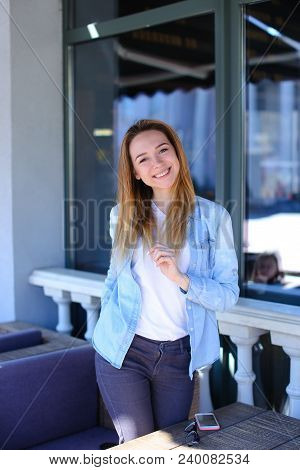 Nice Girl Standing Near Window With Smartphone And Phone On Table. Concept Of Street Cafe And Modern
