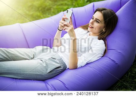 Relax On Fresh Air. Pretty Young Woman Lying On Inflatable Sofa Lamzac Use Mobile Phone While Restin