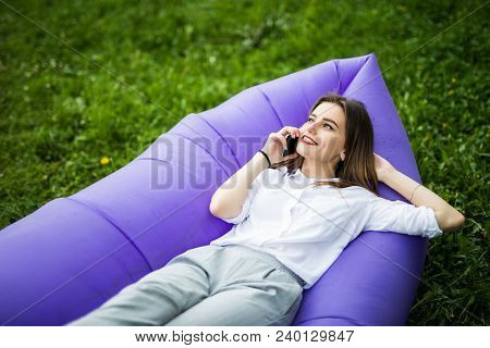 Pretty Young Woman Lying On Inflatable Sofa Lamzak Talk On Phone While Resting On Grass In Park On T