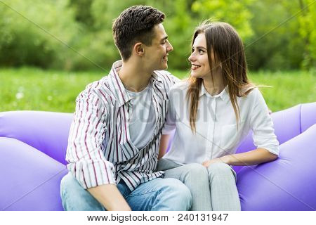 Love Couple Lying On Inflatable Sofa Lamzac On Laptop While Resting On Grass In Park Looking Each Ot