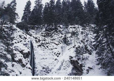Mountain Waterfall With Rocks And Cliffs Covered With Snow During Snowfall, The Mountains Trees