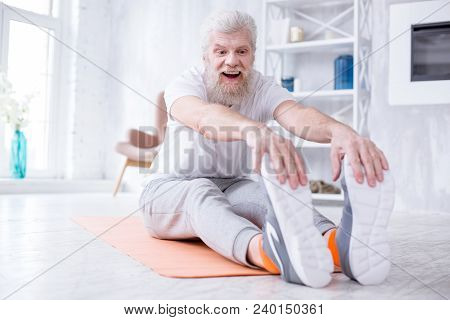 Stretching Muscles. Pleasant Elderly Man Sitting On The Yoga Mat, Tugging At His Toes And Smiling Wi