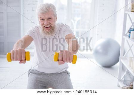 Generating Endorphins. Upbeat Elderly Man Grinning While Exercising With Dumbbells And Doing Squats