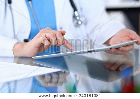 Woman Doctor Using Tablet Computer While Sitting At The Glass Table In Hospital Office, Closeup. Hea