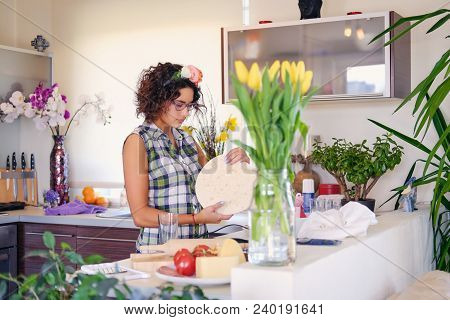 Positive Brunette Female Makes Dough In A Kitchen With Yellow Tulips On Background.