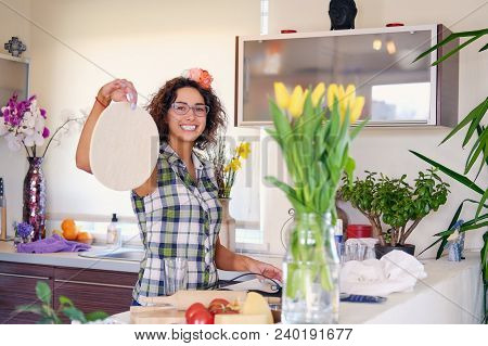 Positive Brunette Female Makes Dough In A Kitchen With Yellow Tulips On Background.