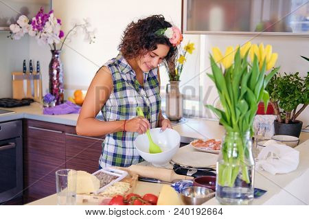 Positive Brunette Female Makes Dough In A Kitchen With Yellow Tulips On Background.