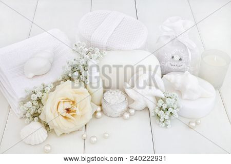 Beauty spa treatment products with a rose flower, ex foliating salt, shell soaps, body lotion, sponges, wash cloths, seashells, gysophelia flowers and decorative pearls on white wood background.

