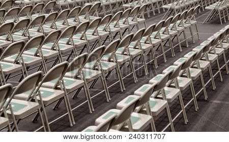 Symmetrical Rows Of Silver Folding Chairs With A Green Folder Placed On The Seat Of Each One