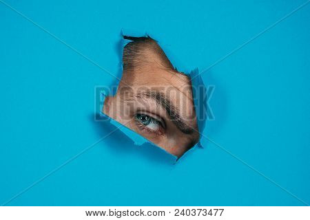 Eye Looking Through Hole In Paper. Spy Eye Watching Through A Hole. Eye Looking Through Hole In Blue